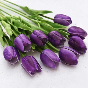 May include: A bouquet of purple artificial tulips with green stems. The tulips are arranged in a fan shape, with the tips of the flowers pointing towards the right side of the image.