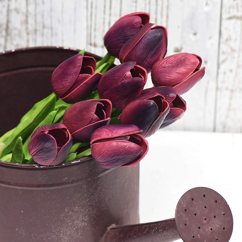 May include: A bouquet of deep burgundy artificial tulips in a brown metal watering can. The tulips are arranged in a cluster, with the stems extending out of the can.