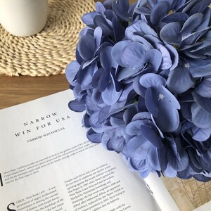 May include: A bouquet of blue hydrangea flowers rests on a wooden surface next to an open book. The book's title reads "NARROW WIN FOR USA". A white ceramic mug and a woven coaster are also visible.