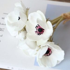 May include: A bouquet of white artificial anemones with pink and black centers. The flowers are arranged in a cluster and have long green stems.