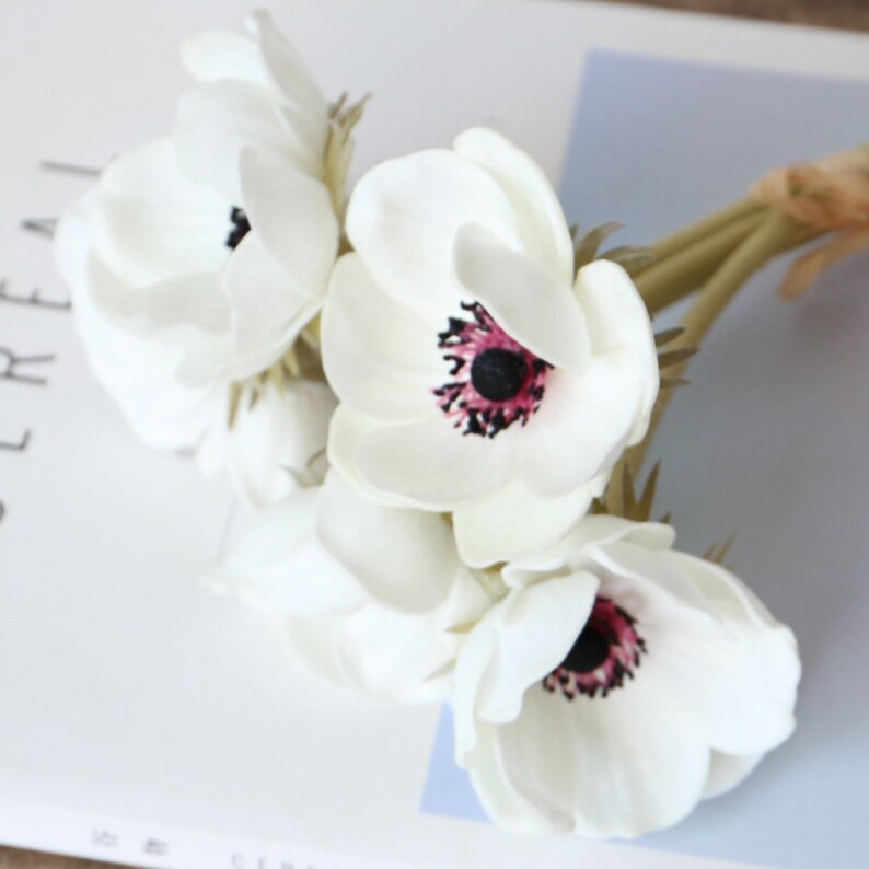May include: A bouquet of white artificial anemones with pink and black centers. The flowers are arranged in a cluster and have long green stems.