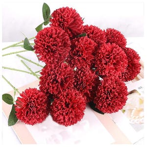 May include: A bundle of artificial red dahlia flowers with green stems and leaves. The flowers have a dense, textured appearance, and the stems are long and slender. The arrangement is set against a white background.
