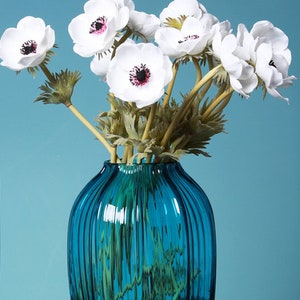 May include: A bouquet of white anemones with dark centers arranged in a blue glass vase with vertical ridges. The vase is on a black surface.