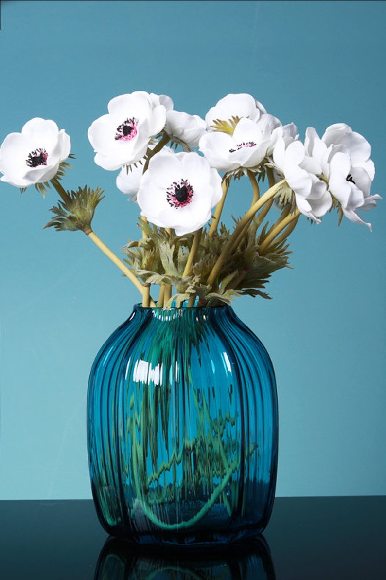 May include: A bouquet of white anemones with dark centers arranged in a blue glass vase with vertical ridges. The vase is on a black surface.