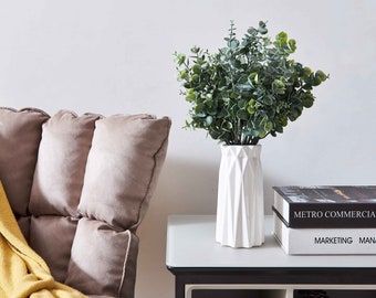 May include: A white ceramic vase with a geometric design holds a bouquet of green eucalyptus leaves. The vase is on a black side table with a glass top. Two books are stacked on the table, one with the text "METRO COMMERCIA" and the other with the text "MARKETING MANA".