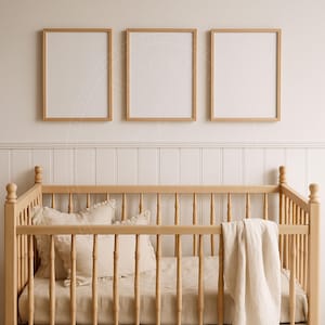 May include: A light wood baby crib with a cream-colored blanket and pillows. Three framed prints with light wood frames hang above the crib. The wall behind the crib is white with vertical paneling.