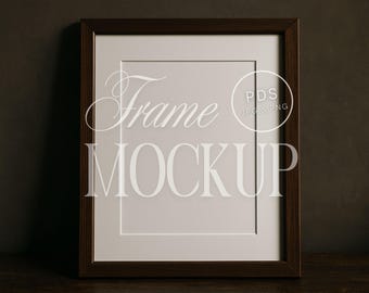 May include: A dark brown wooden frame mockup with a white mat and a blank white space for an image or artwork. The words "Frame MOCKUP" are displayed in a stylized font. The frame is on a dark wooden surface.
