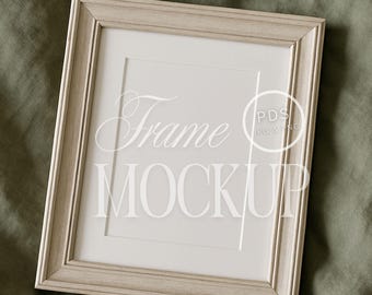 Może przedstawiać: Makieta ramki z jasnego drewna z białą passe-partout i białym tłem. Ramka leży na zielonej powierzchni z tkaniny. Tekst "Frame Mockup" jest widoczny w środku ramki.
