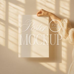 May include: A white poster mockup with the text "Poster Mockup" in a script font. The poster is held up by a hand against a beige wall with light shining through a window, creating shadows.