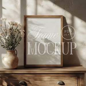 May include: A wooden frame mockup with a white background, sitting on a wooden dresser with a vase of white flowers. The text "Frame Mockup" is written in white on the frame.