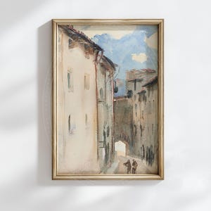 May include: Watercolor painting of a European street scene in a gold frame. The artwork depicts buildings with arched doorways and a blue sky. Two figures are walking down the street. The painting has a vintage aesthetic.