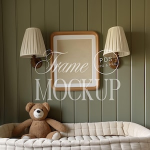 May include: A light brown teddy bear sits on a white cushioned bench in front of a green wall. A wooden frame with a blank white mat is hanging on the wall. Two white lampshades with wooden bases are mounted on the wall on either side of the frame.
