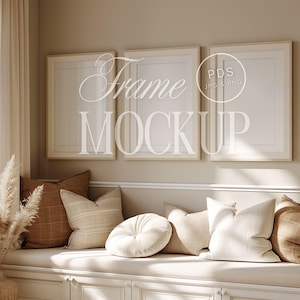 May include: Three white picture frames with a cream colored mat are hanging on a beige wall above a white bench with brown and cream pillows. The frames are empty and ready for your photos or artwork.