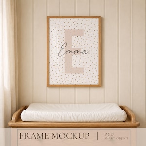 May include: A rectangular wooden frame containing a print with a light beige background, gold polka dots, a large light beige letter E, and the name Emma written in black cursive script. The frame is mounted on a vertical paneled wall.