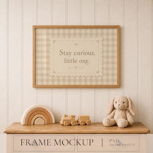 May include: A rectangular wooden frame containing a beige print with a light brown gingham pattern and the text Stay curious, little one. Below the frame sits a wooden rainbow stacker, a wooden toy train, and a beige plush rabbit toy.