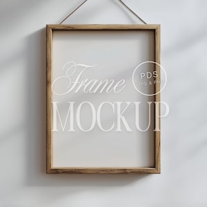 Puede incluir: Un marco de madera marrón con fondo blanco. El marco está colgado en una pared blanca con una sombra de una ventana. El texto "Frame Mockup" está escrito en blanco en el marco. El texto "PDS JPG & PNG" está escrito en blanco en un círculo en el marco.
