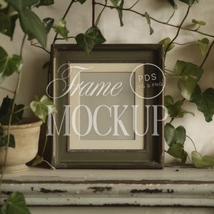May include: A green picture frame with a white mount and a blank space for a photograph. The frame is sitting on a white shelf with green ivy plants on either side.