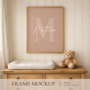 May include: A rectangular wooden frame containing a dusty pink print with a large, light-colored letter M and the name Matteo written in white cursive script. The frame is displayed on a wooden dresser next to a small, light brown teddy bear.