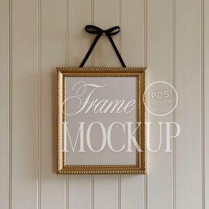 Könnte beinhalten: Ein goldener Bilderrahmen mit einer Perlenbordüre hängt an einem schwarzen Band vor einem weißen Holzpaneel-Hintergrund. Der Rahmen ist leer und bereit für ein Foto oder ein Kunstwerk. Der Text "Frame Mockup" ist in Weiß auf den Rahmen gedruckt.