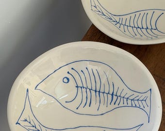 May include: Two round, off-white ceramic bowls featuring hand-painted blue line art of fish skeletons. Each bowl displays two fish motifs oriented in opposite directions, rendered in a simple, minimalist style with visible brush strokes.