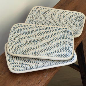 May include: Two rectangular ceramic trays with rounded corners, featuring an off-white base colour decorated with a dense pattern of small, hand-painted blue dots distributed across the entire surface of each piece.