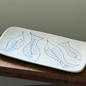 Puede incluir: Una bandeja de servicio rectangular de cerámica blanca con cuatro esqueletos de pescado azules dibujados a mano en fila. La bandeja tiene bordes ligeramente curvados y un acabado liso y brillante con un diseño de líneas.