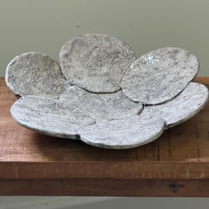 Peut inclure: Un bol en céramique peu profond en forme de fleur avec une finition texturée et tachetée blanc cassé et gris. Le bol est composé de plusieurs segments arrondis et superposés en forme de pétales formant un bassin circulaire irrégulier.