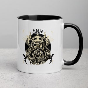 May include: White ceramic mug with a black rim and handle. The mug features a black and white illustration of Odin, a Norse god, with the word "ODIN" written above him. The illustration is surrounded by lightning bolts and runes.