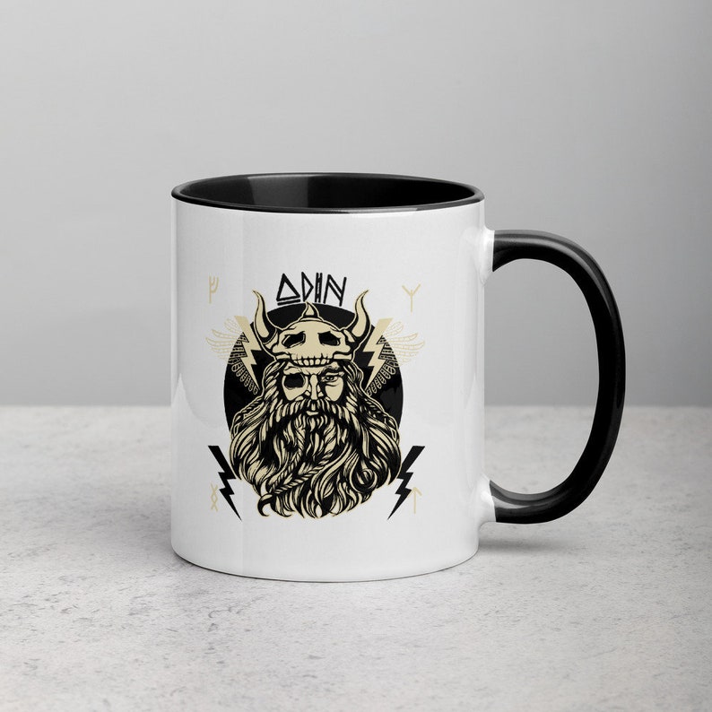 May include: White ceramic mug with a black rim and handle. The mug features a black and white illustration of Odin, a Norse god, with the word "ODIN" written above him. The illustration is surrounded by lightning bolts and runes.