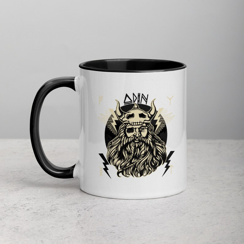 May include: White ceramic mug with a black rim and handle. The mug features a black and white illustration of Odin, a Norse god, with the text "ODIN" above him.