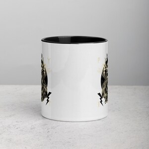 May include: White ceramic mug with a black rim and handle. The mug features a black and gold illustration of a winged creature with a crown and lightning bolts. The illustration is repeated on both sides of the mug.