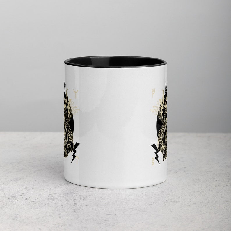 May include: White ceramic mug with a black rim and handle. The mug features a black and gold illustration of a winged creature with a crown and lightning bolts. The illustration is repeated on both sides of the mug.