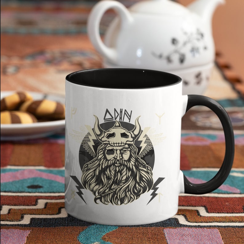 May include: White ceramic mug with a black rim and handle. The mug features a black and white illustration of a Viking warrior with a beard and horns. The text "Odin" is printed above the warrior's head.