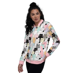 Puede incluir: Una chaqueta bomber blanca con una cremallera rosa y un estampado de gatos coloridos por todas partes. Los gatos están en varios colores y poses.
