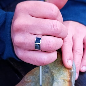 Puede incluir: Un anillo de plata con una piedra cuadrada negra engastada en el centro. El anillo está en el dedo de una persona.