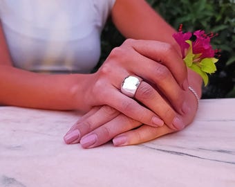 May include: A silver ring with a large, rounded, square-shaped top. The ring is on a person's finger.