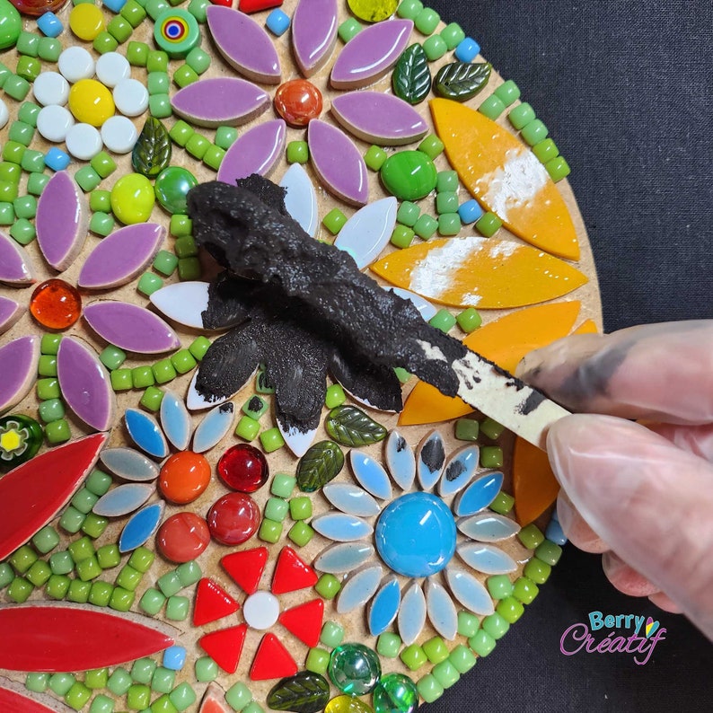 Bright flower mosaic tiles are part covered in a black grout, the wooden mixing stick is spreading the grout across the surface of the mosaic kit