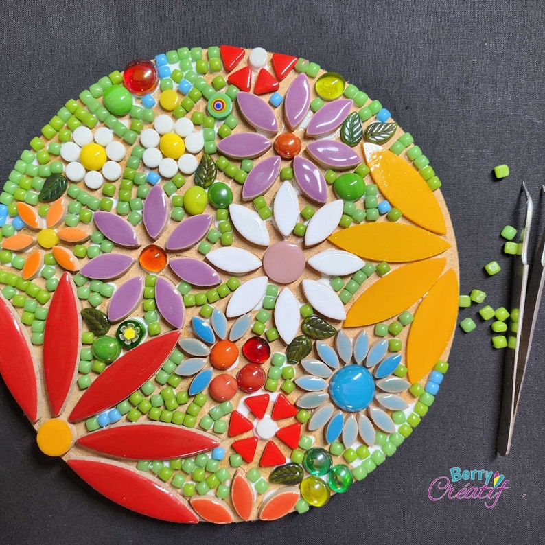 Work in progress of a flower mosaic kit, with all of the colorful tiles stuck to the circular mdf base. The board is sitting on a black background and there are metal tweezers and an odd remaining mosaic tile around the edge.
