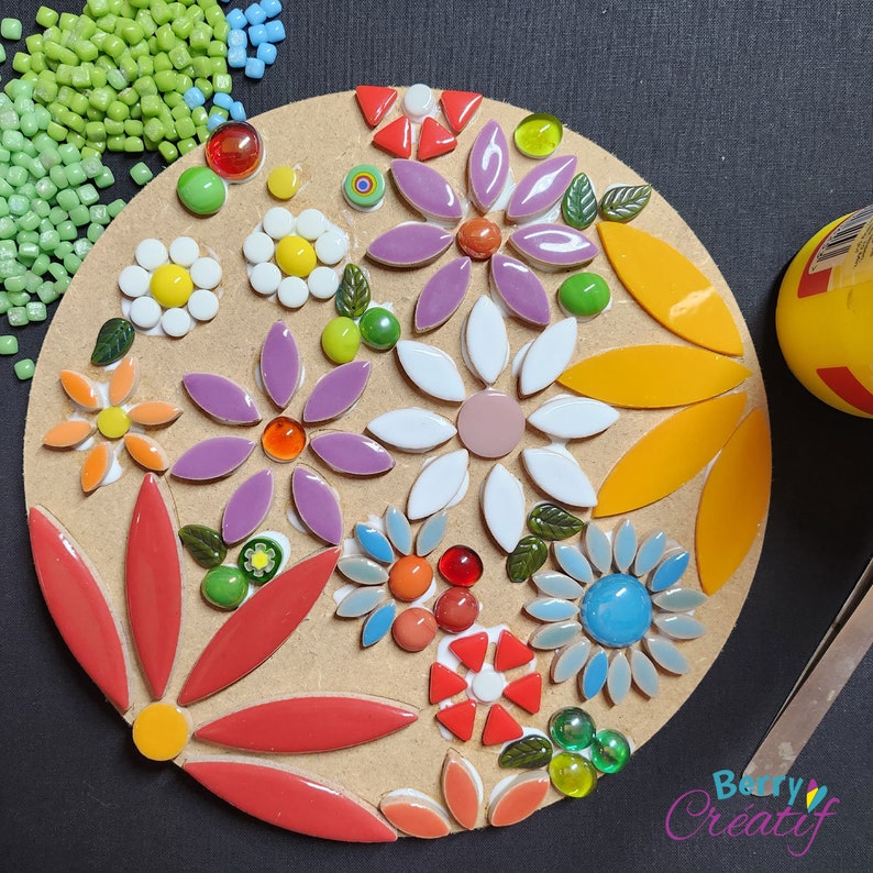 Work in progress of a flower mosaic kit, with some of the colorful tiles stuck to the round mdf base. The board is sitting on a black background and there are some loose mosaic tiles, tweezers and glue around the edge.
