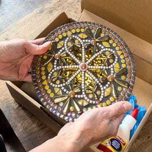Peut inclure: Une pièce d'art en mosaïque ronde avec un motif de libellule dans les tons or, jaune et marron. La mosaïque est présentée dans une boîte en carton, mettant en valeur les détails complexes et le savoir-faire de l'œuvre. La pièce mesure environ 20 cm de diamètre.
