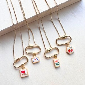 May include: Four gold-toned necklaces featuring thin box chains and oval carabiner clasps. Each necklace has a small, square or hexagonal pendant with colorful enamel illustrations of a red flower, a letter, a pink flower, and two strawberries.