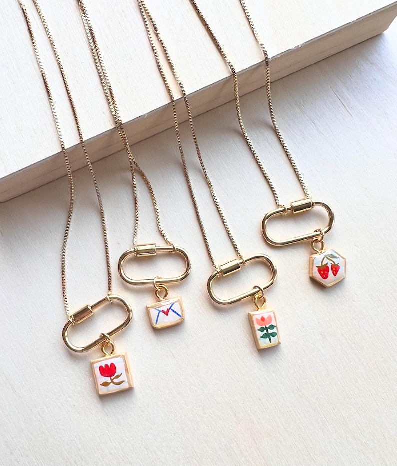 May include: Four gold-toned necklaces featuring thin box chains and oval carabiner clasps. Each necklace has a small, square or hexagonal pendant with colorful enamel illustrations of a red flower, a letter, a pink flower, and two strawberries.