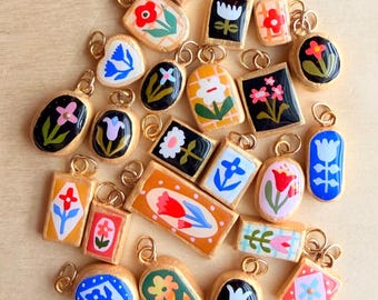 May include: A collection of small, assorted metal-rimmed charms featuring hand-painted floral designs in various shapes including rectangles, ovals, and arches. Each charm includes a gold-toned jump ring for attachment, showcasing vibrant colors.