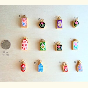 May include: A collection of twelve small gold-rimmed enamel charms featuring various floral and house designs, numbered one through twelve, displayed alongside a US dime for scale. Each charm has a small loop for attachment to jewelry or accessories.