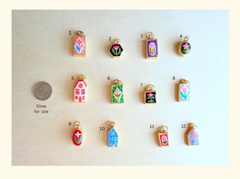 May include: A collection of twelve small gold-rimmed enamel charms featuring various floral and house designs, numbered one through twelve, displayed alongside a US dime for scale. Each charm has a small loop for attachment to jewelry or accessories.