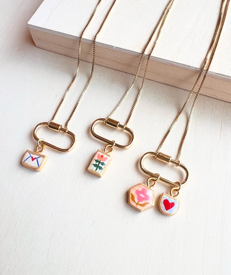 May include: Three gold-toned necklaces featuring oval carabiner-style clasps. The first pendant is a white envelope with a red heart. The second is a rectangular floral charm. The third features a hexagonal pink flower charm and a small heart charm.