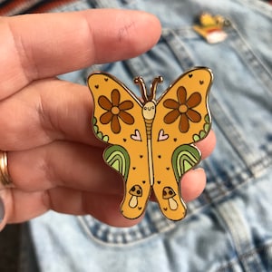 Könnte beinhalten: Eine gelbe Schmetterlings-Emaille-Anstecknadel mit braunen Blumen, grünen Wirbeln und rosa Herzen. Der Schmetterling hat ein süßes Gesicht mit zwei Augen und einem kleinen Lächeln. Am unteren Ende des Schmetterlings befinden sich zwei kleine Pilze.