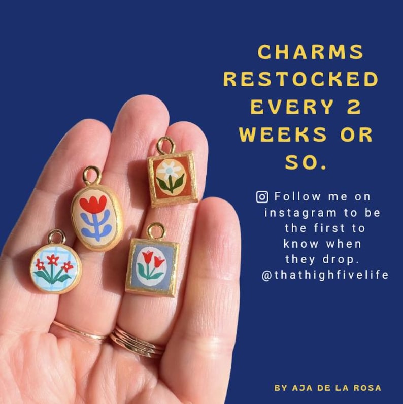 May include: Four small gold-toned charms with hand-painted floral designs in red, blue, and white. The charms are round and square-shaped, each featuring a small loop at the top. Text on the right reads: CHARMS RESTOCKED EVERY 2 WEEKS OR SO.