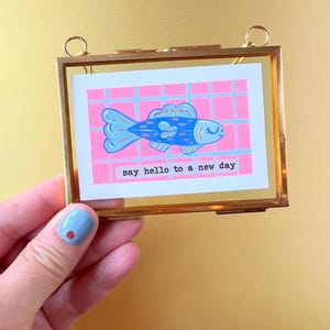 Könnte beinhalten: Ein gerahmter Druck mit einer blauen Fischillustration auf einem rosa Gitterhintergrund. Der Druck enthält den Text "say hello to a new day" in weißer Schrift. Der Rahmen ist goldfarben mit einer transparenten Glasfront.