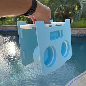 Op de afbeelding: Een lichtblauwe plastic pool float houder met twee ronde bekerhouders en een centrale cilindrische sectie. De houder wordt door een hand omhoog gehouden. De achtergrond is een zwembad.